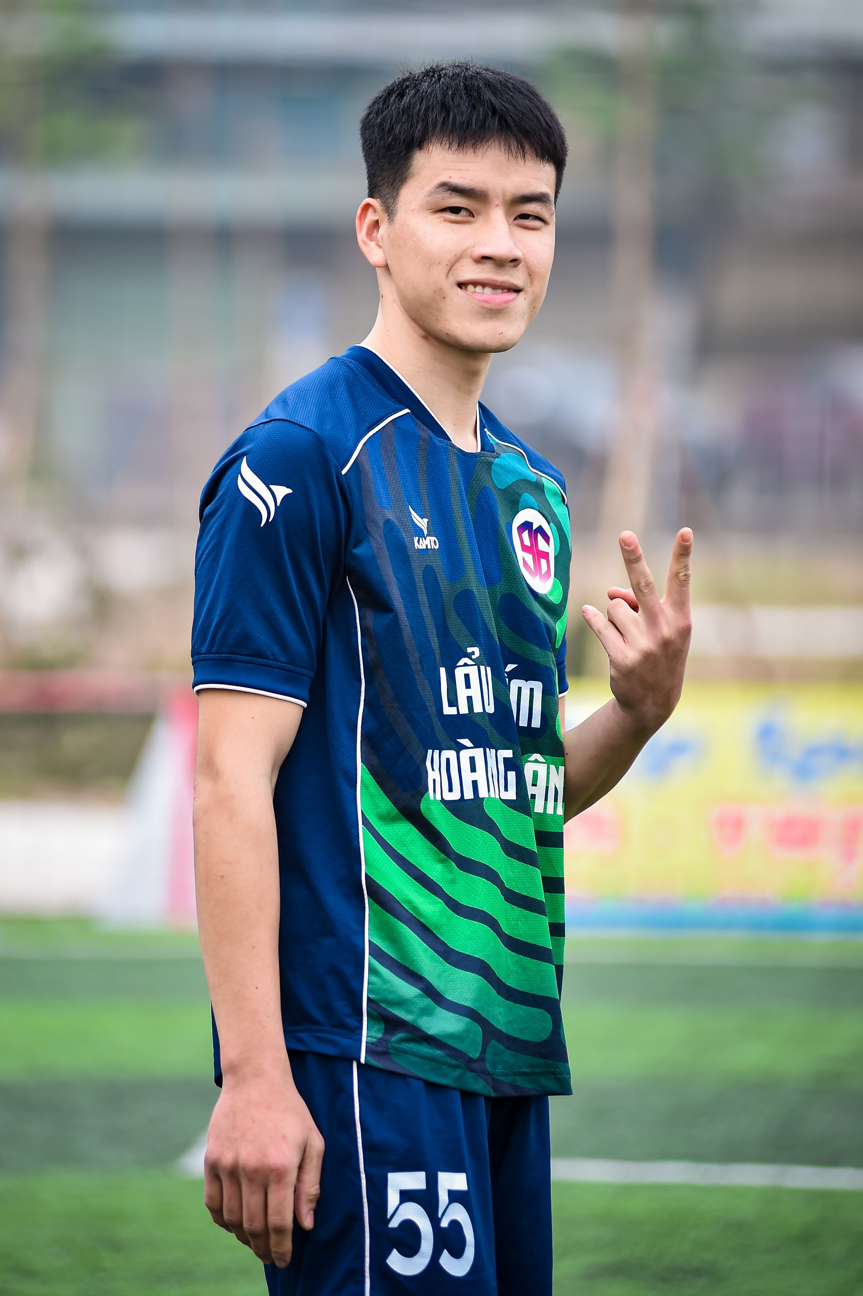 Young football player in blue uniform smiling and gesturing peace sign outdoors in Hà Nội, Vietnam.