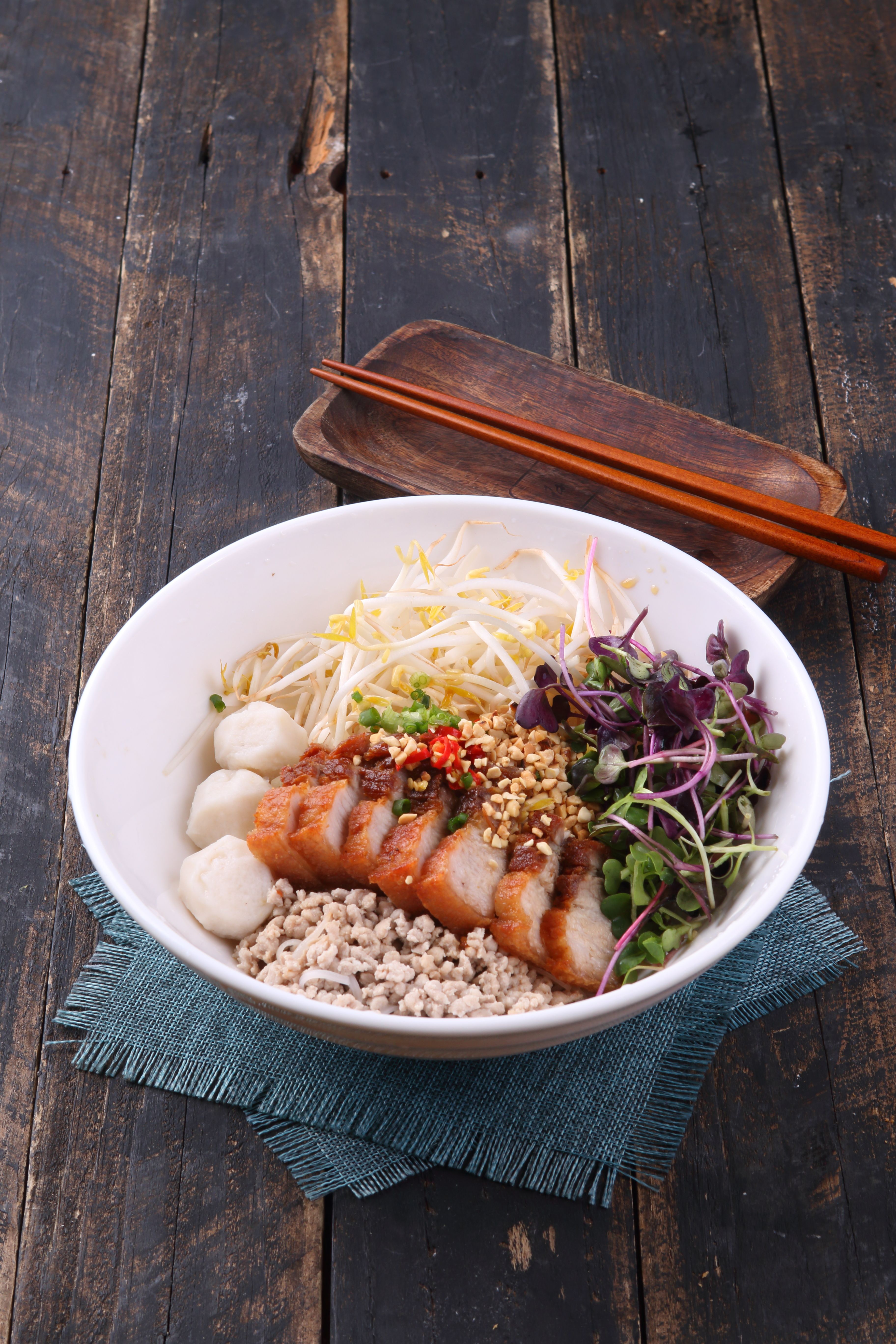 Asian noodle bowl with pork, vegetables, and herbs on a rustic wooden table.