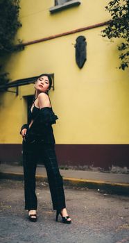 Fashionable woman in elegant outfit poses confidently against a yellow wall in an urban setting.