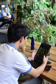 Focused young man using a tablet at an outdoor cafe surrounded by greenery and tech devices.