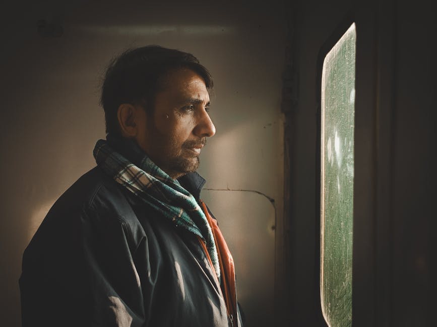 A man with a scarf stares pensively out of a train window, captured indoors.