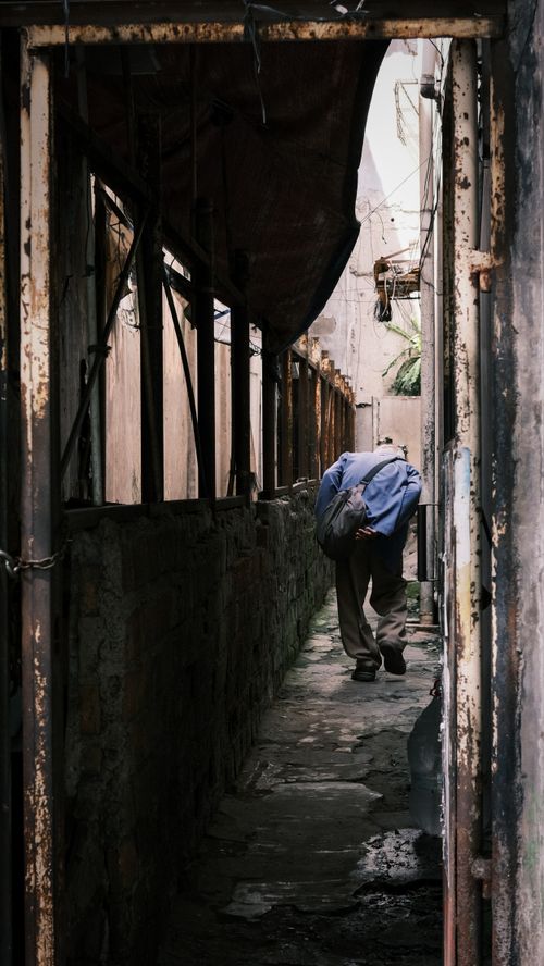 Free A senior man in a blue jacket walks down a narrow, rustic alleyway, evoking solitude. Stock Photo