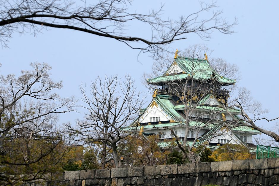 A view of Osaka Castle from behind bare trees, showcasing its historical architecture against a winter sky.