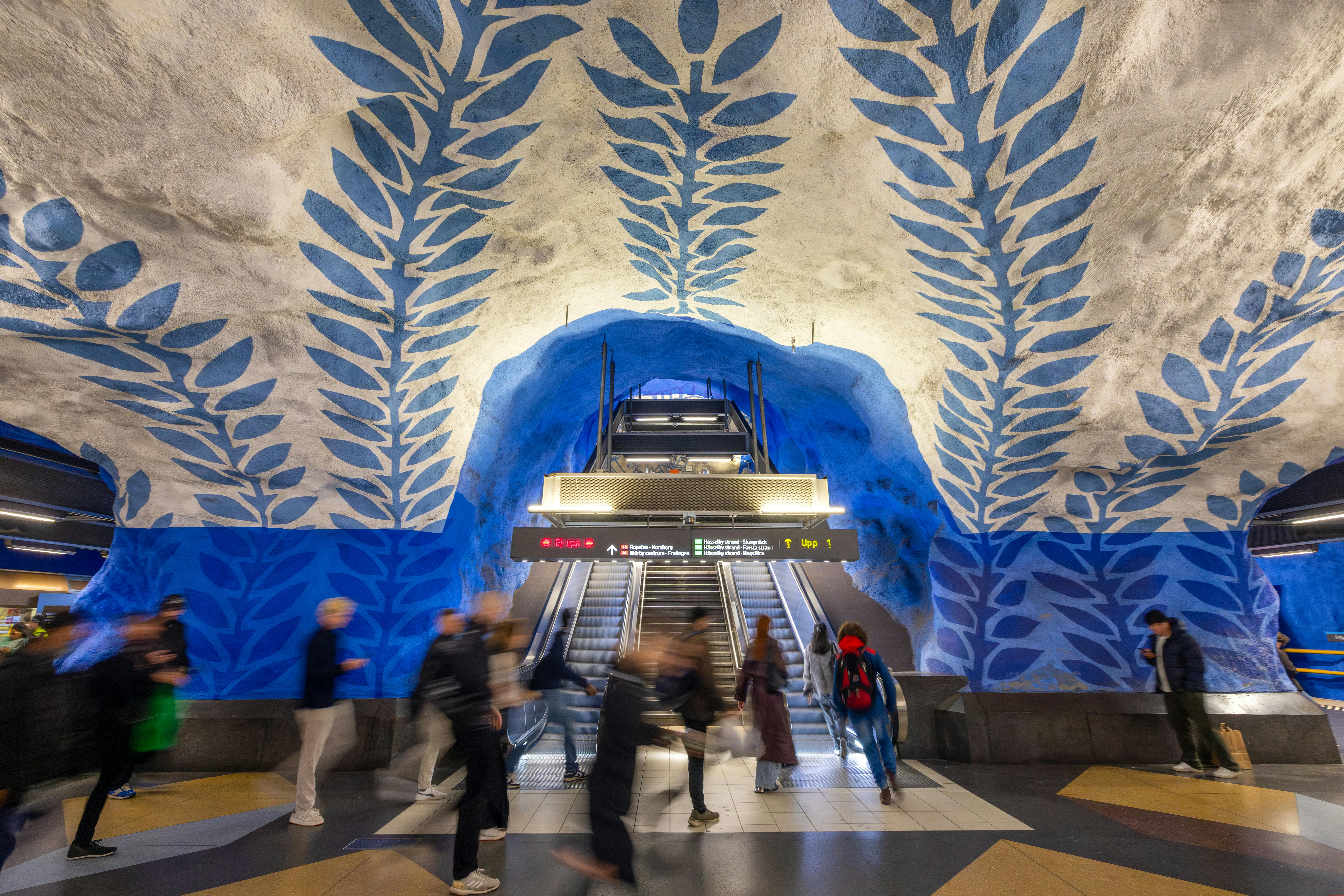 Blurred motion of people at an artistic Stockholm metro station with blue leaf ceiling.