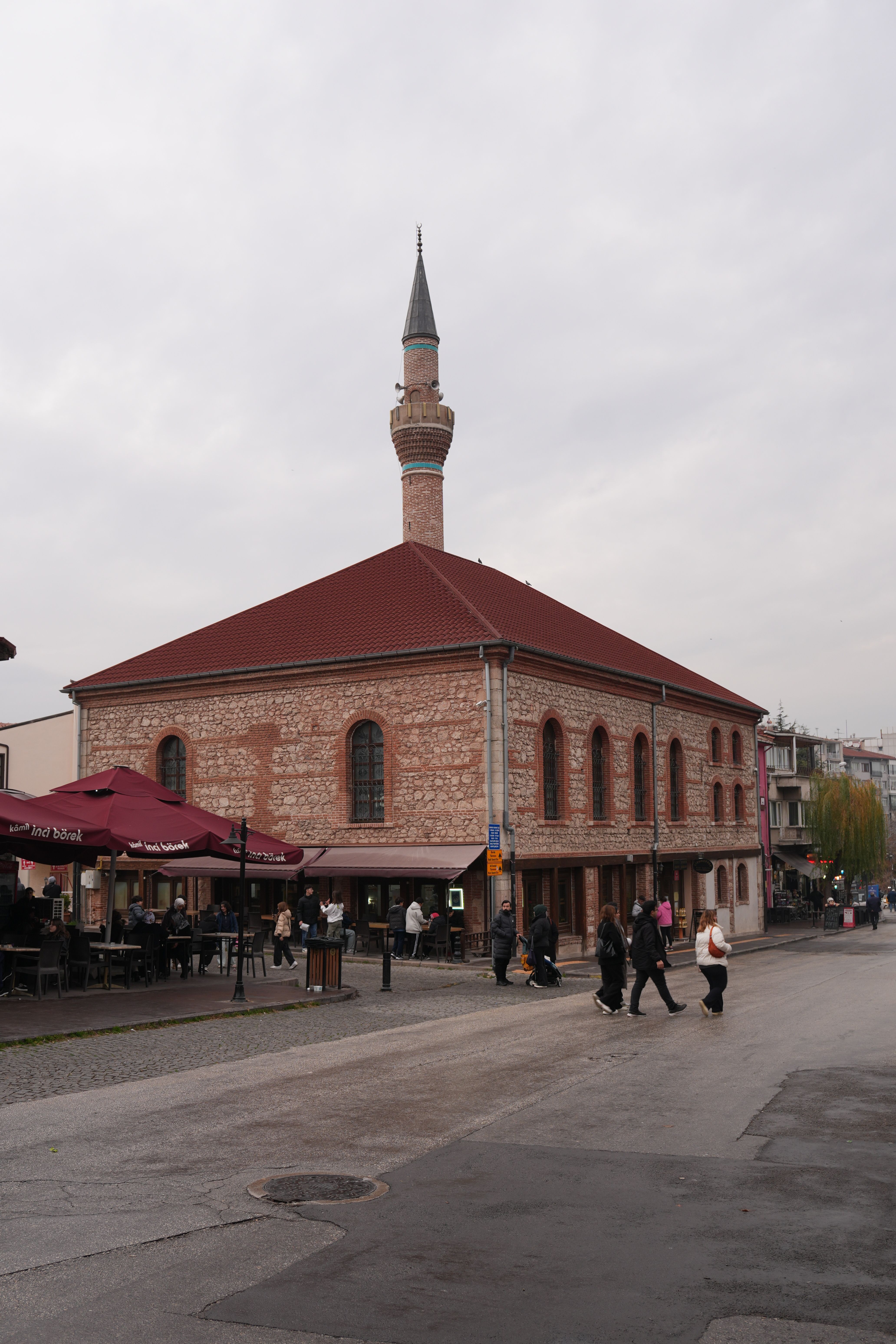 Beautiful historic mosque with red roof and minaret in a bustling urban area, people walking by.