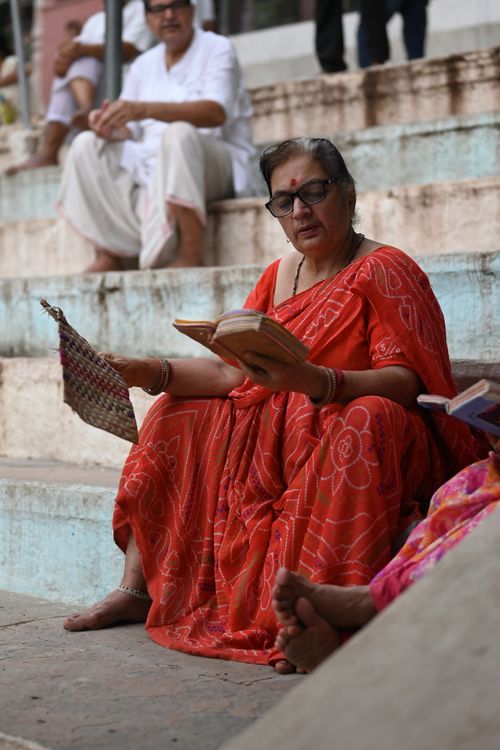 Free A senior woman in a vibrant red saree reads on temple steps, depicting a serene cultural moment in India. Stock Photo