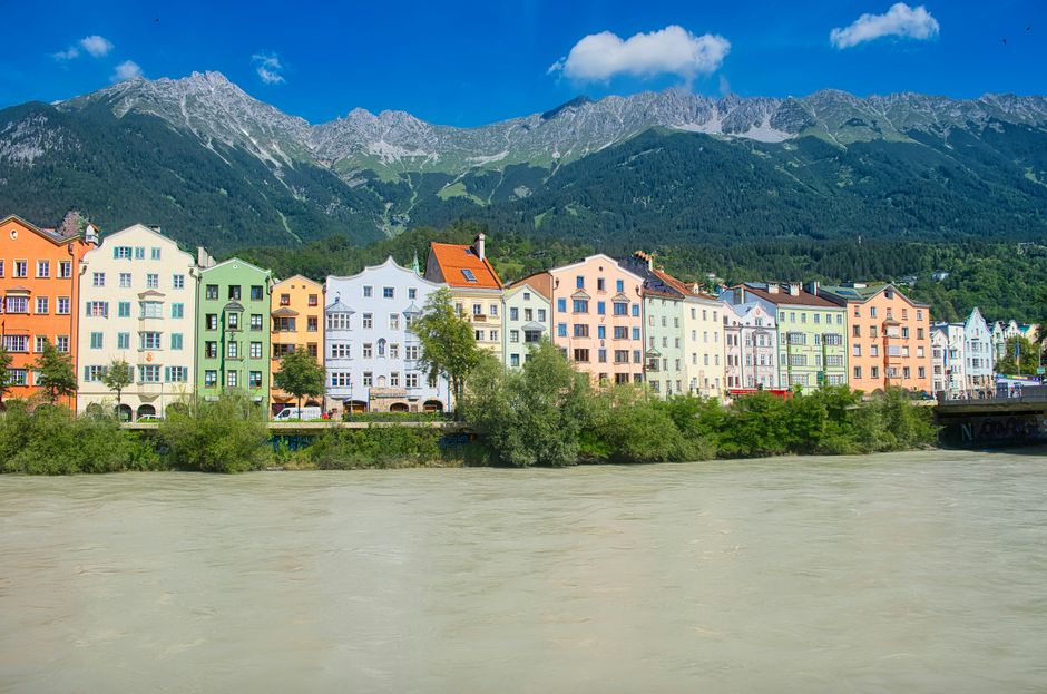 Brightly colored houses along Innsbruck river with mountain backdrop under clear blue sky.