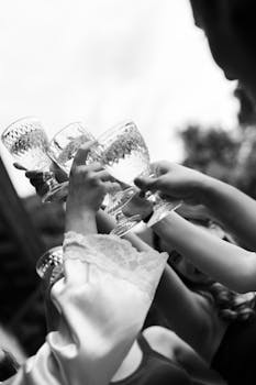 A joyful bridal party raises crystal glasses in a wedding toast, captured in black and white.