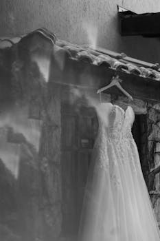 A black and white photo of a beautiful bridal gown hanging outdoors on a rustic wall, showing delicate lace details.