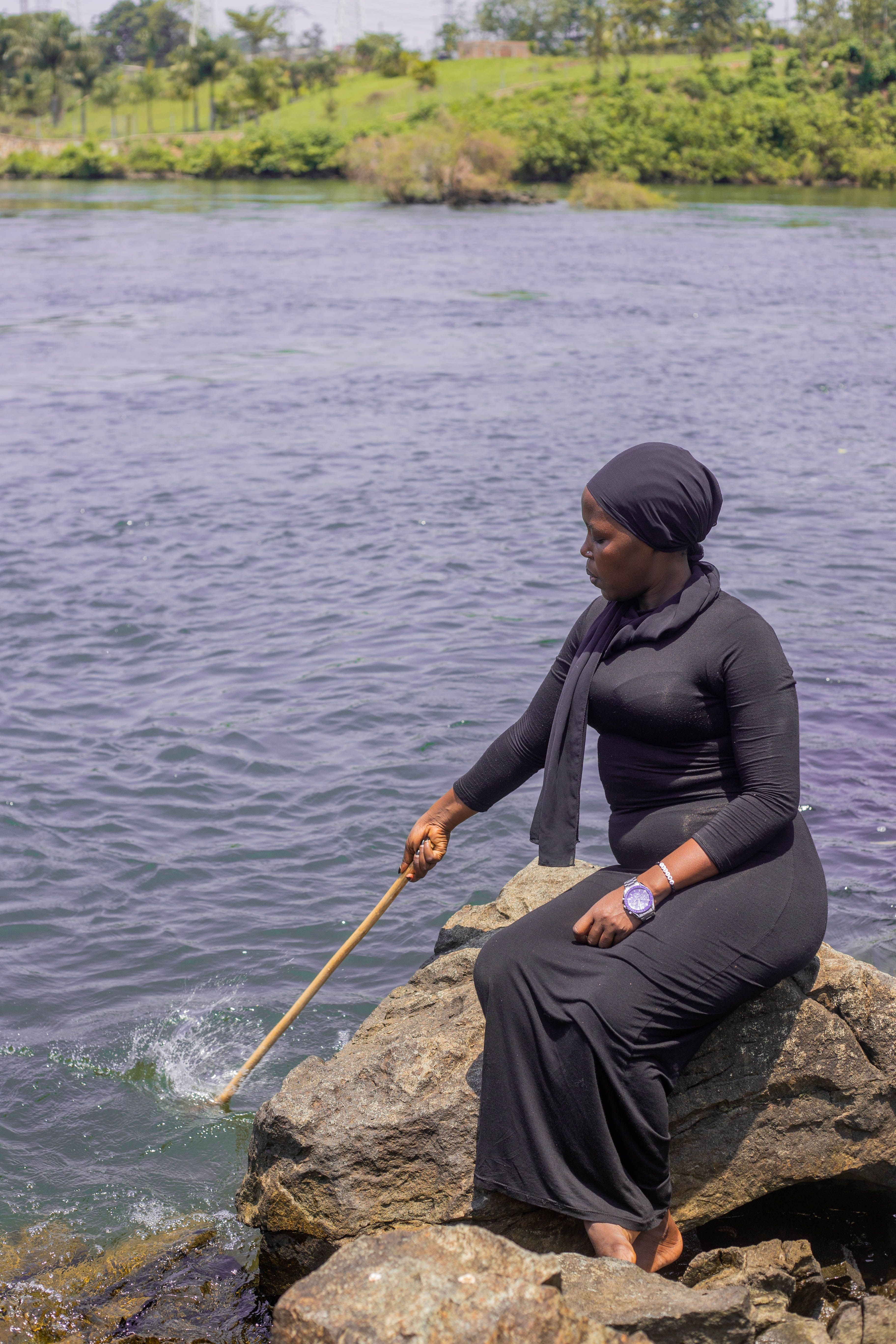 Woman in black dress sitting on rocks by a river using a stick. Peaceful outdoor setting.