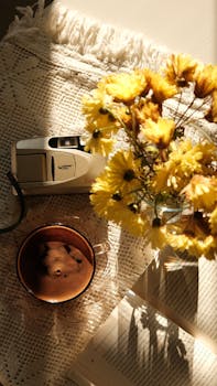 A vintage camera beside yellow flowers and coffee on a sunlit cloth.