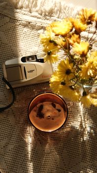 A vintage camera, coffee, and flowers on a textured cloth with sunlight.