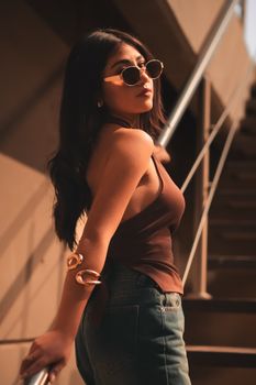 Fashionable woman in sunglasses poses on an outdoor staircase in warm lighting.