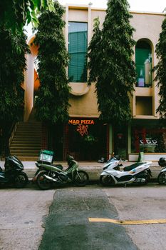 A lively street scene in Bangkok, Thailand featuring motorbikes parked in front of a modern pizza restaurant.
