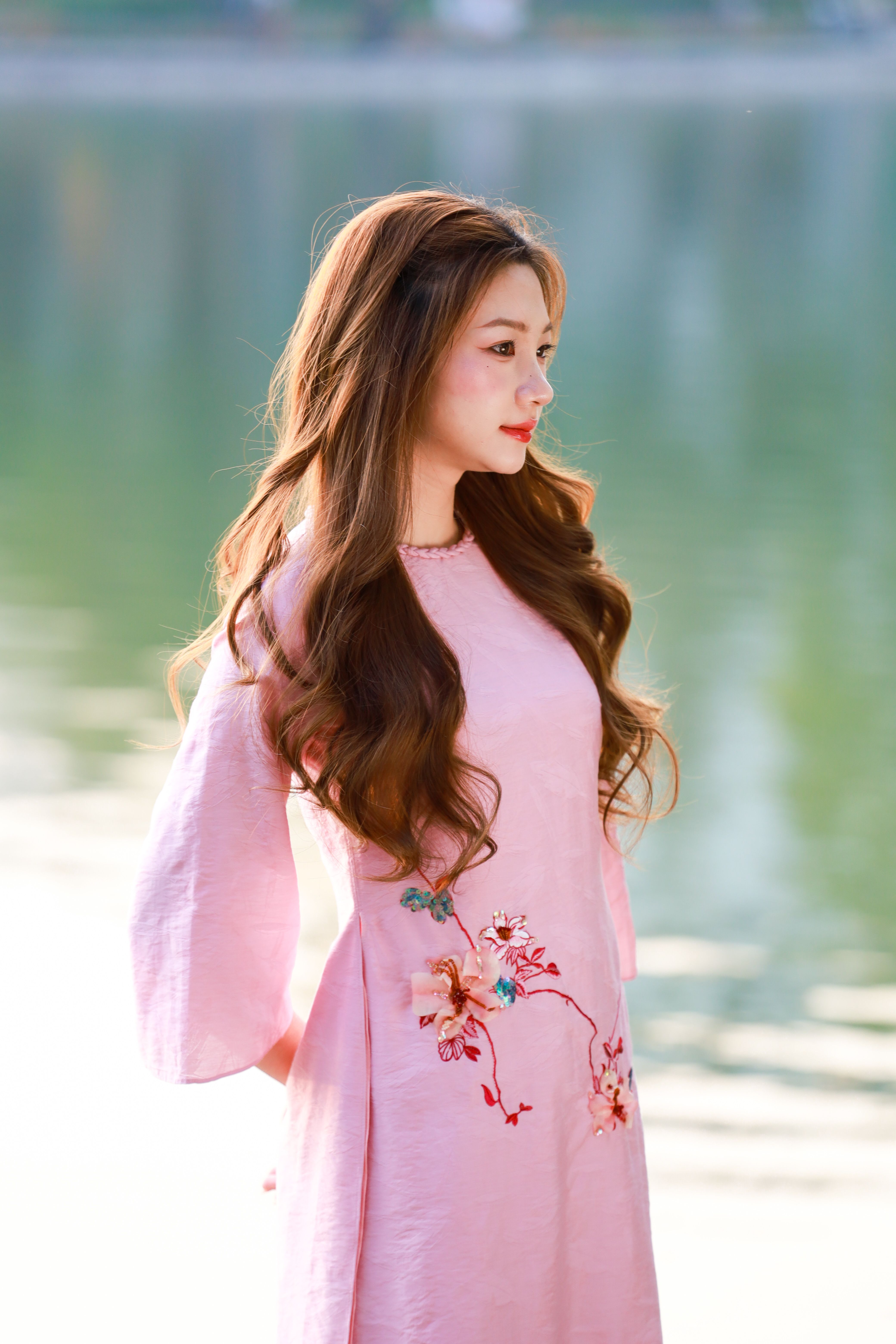 A serene portrait of a woman in a pink traditional dress by a lake in Hà Nội, Việt Nam.
