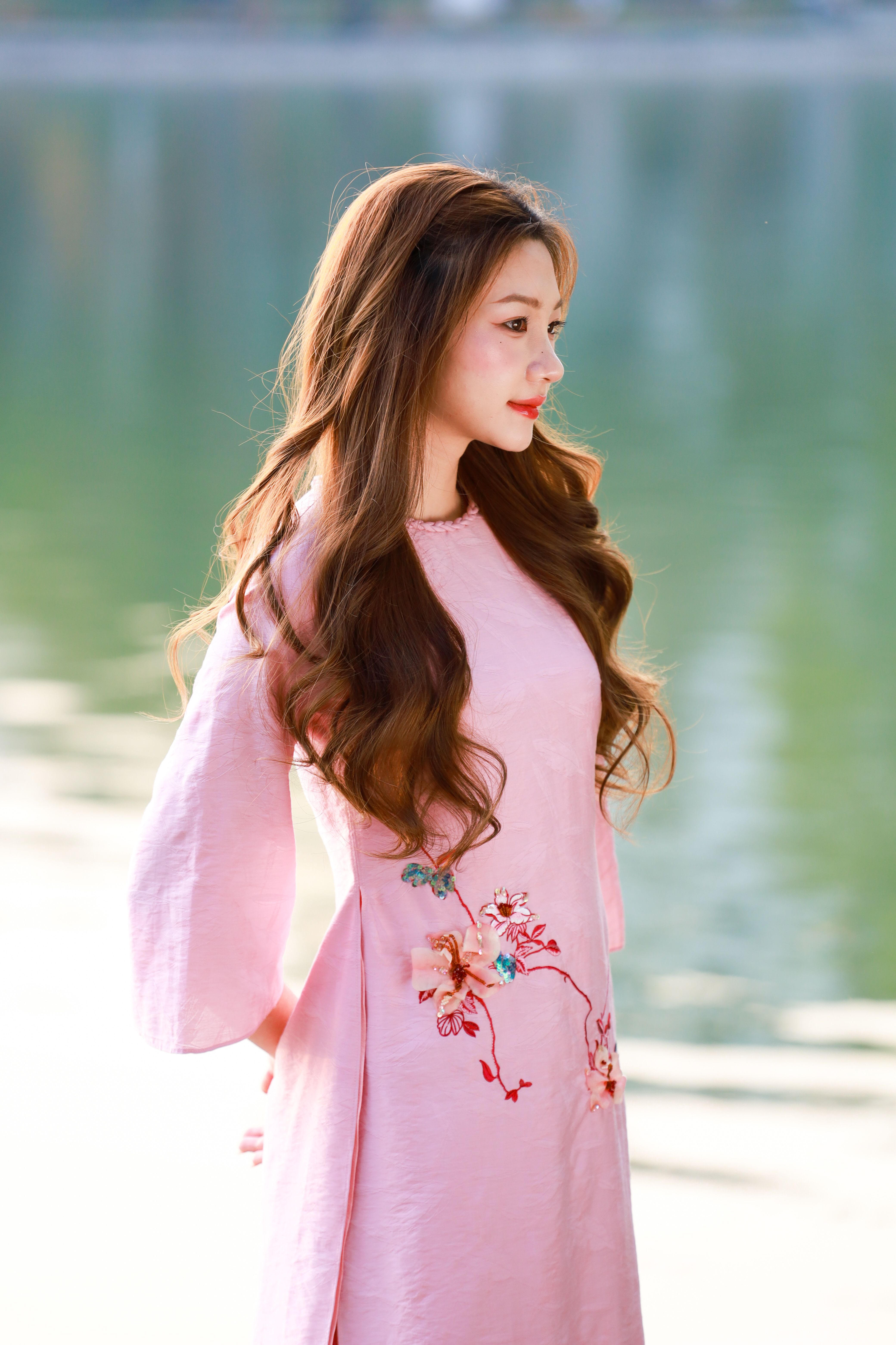 Elegant woman in pink ao dai dress posing by a serene lake in Hanoi, Vietnam.