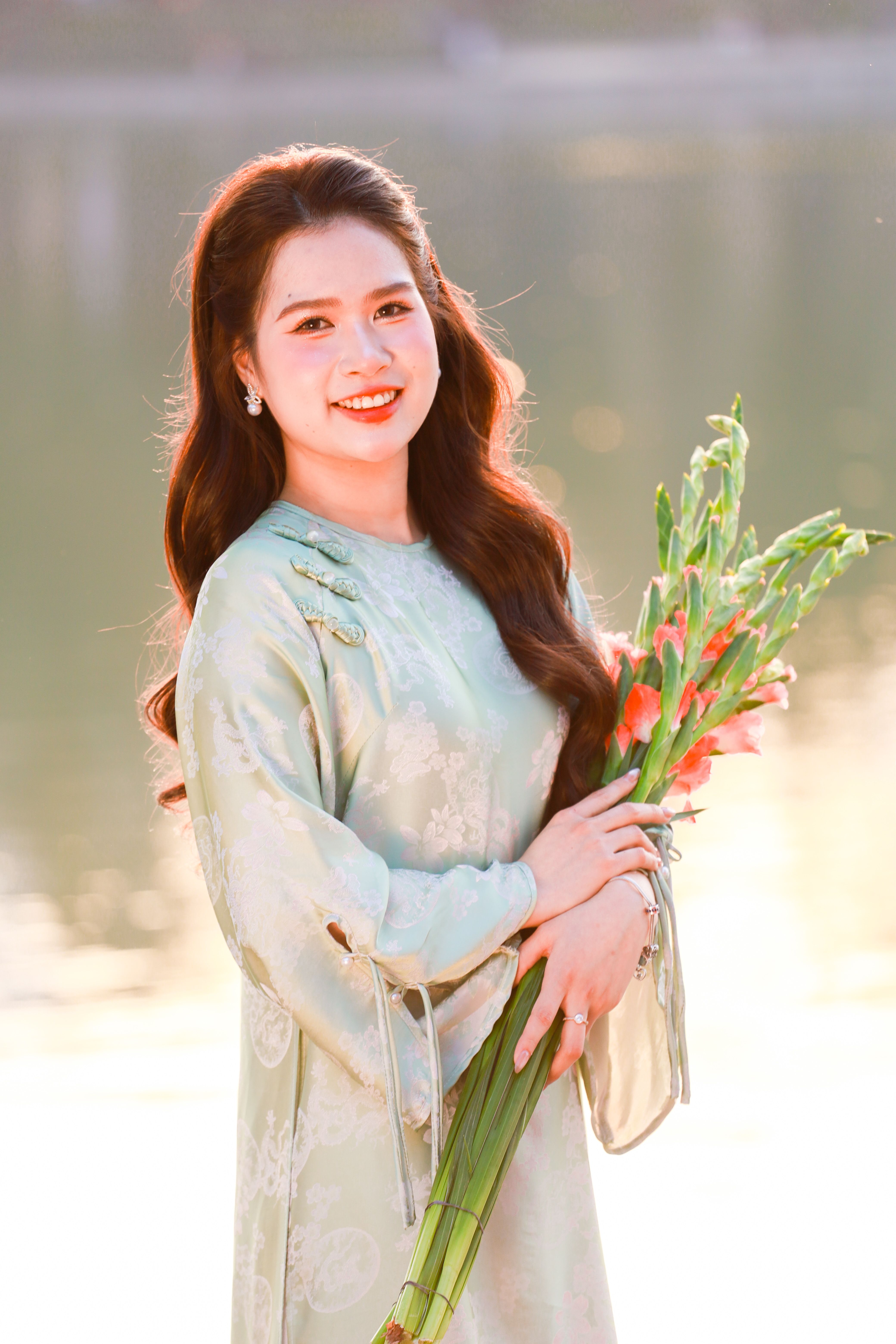 Portrait of a young woman in traditional Vietnamese dress holding flowers, smiling outdoors.