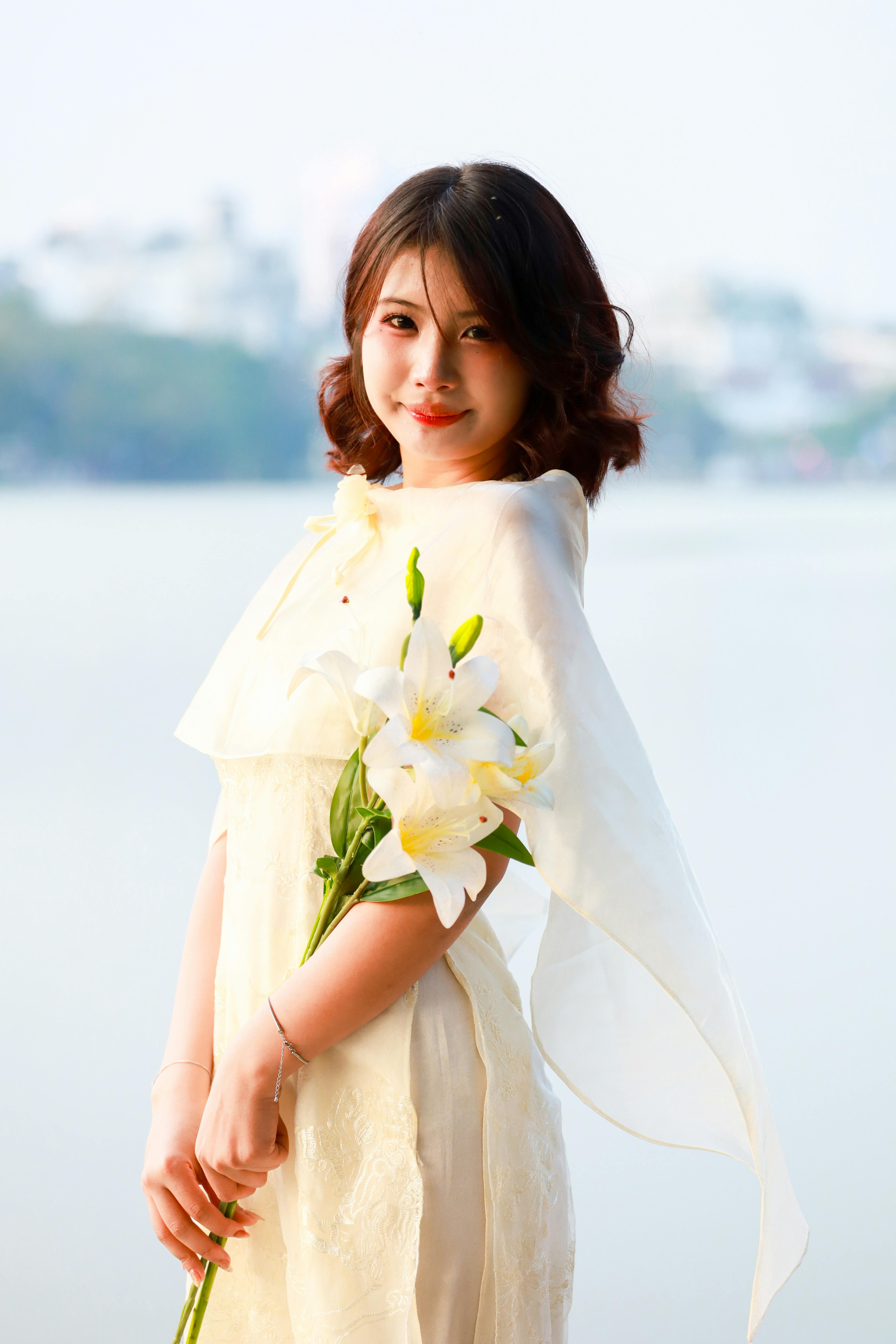 Elegant woman in a long dress holding lilies beside a lake in Hanoi, Vietnam.