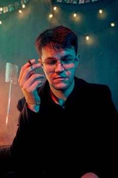 A man in glasses holds a cigarette indoors under warm lighting, creating a moody atmosphere.
