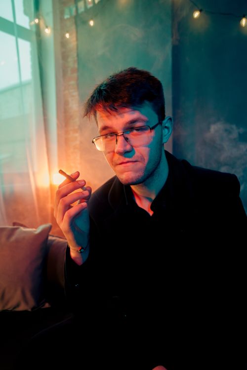 Free Man in glasses holding cigarette in a dimly lit room with a moody atmosphere. Stock Photo