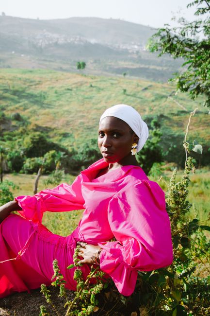 Woman in vibrant pink dress with headscarf posing outdoors in lush landscape.