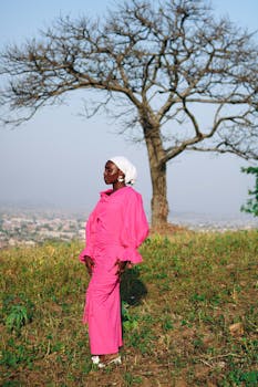 Elegant woman in vibrant pink dress posing outdoors with a scenic view in Abuja, Nigeria.