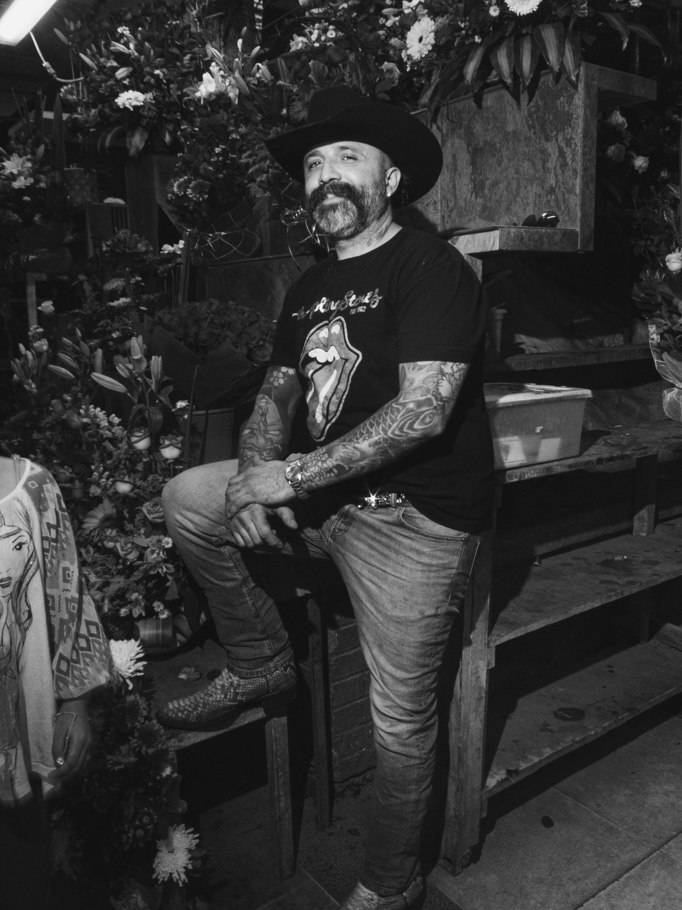 Black and white portrait of a tattooed man wearing a hat, sitting in a flower market in León, Mexico.