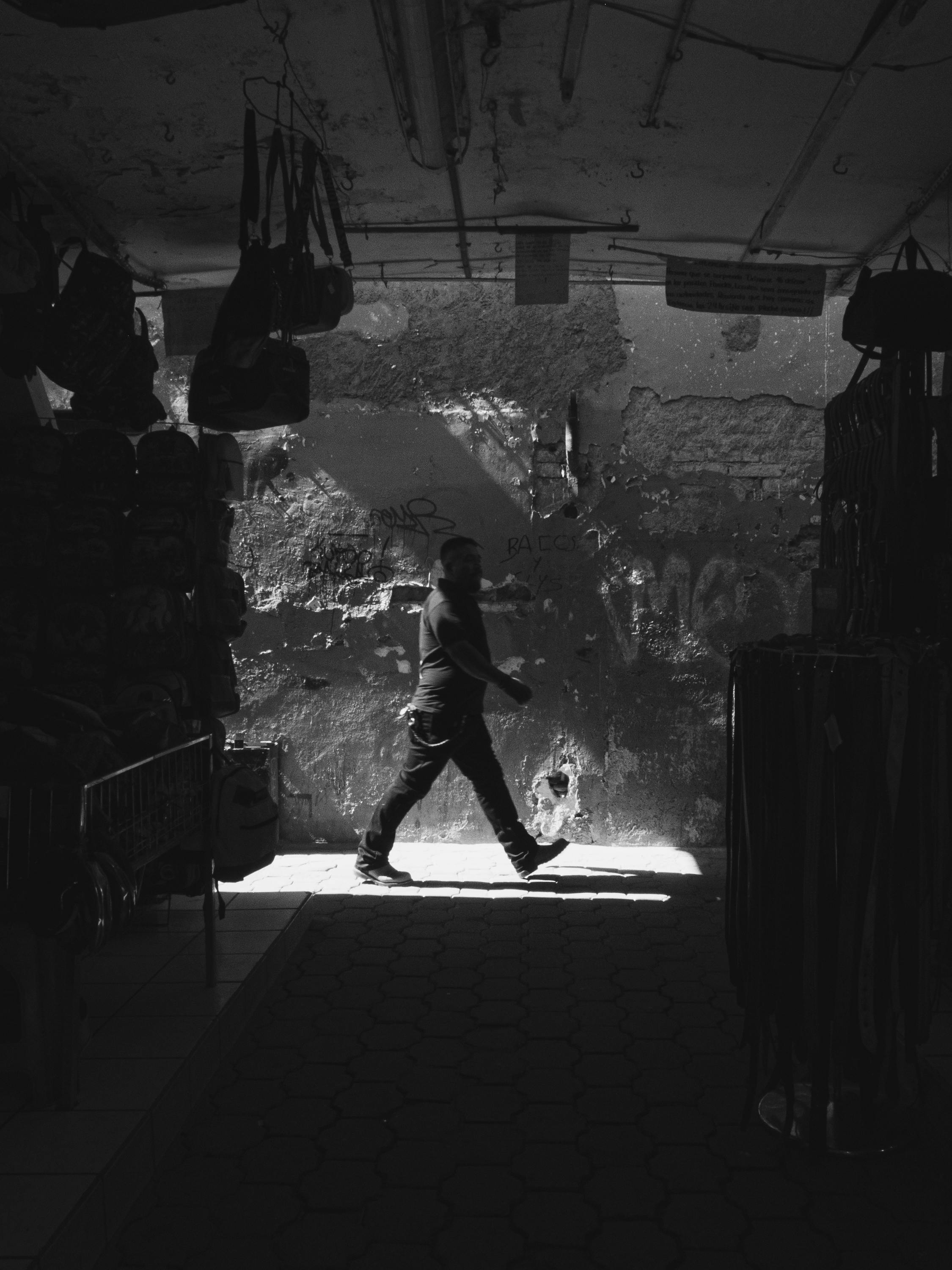 Black and white street photo of man walking in León de los Aldama market, Mexico, highlighting contrast and light.