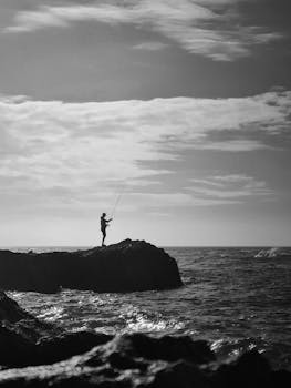 A man fishing on rocky cliffs by the ocean in Morocco, captured in black and white.