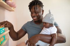 A joyful father with dreadlocks holding his baby indoors, selecting a book.