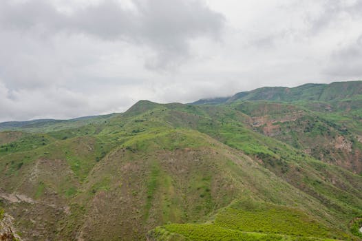 Expansive view of green hills and mountains under a dramatic cloudy sky, showcasing natural beauty and tranquility.