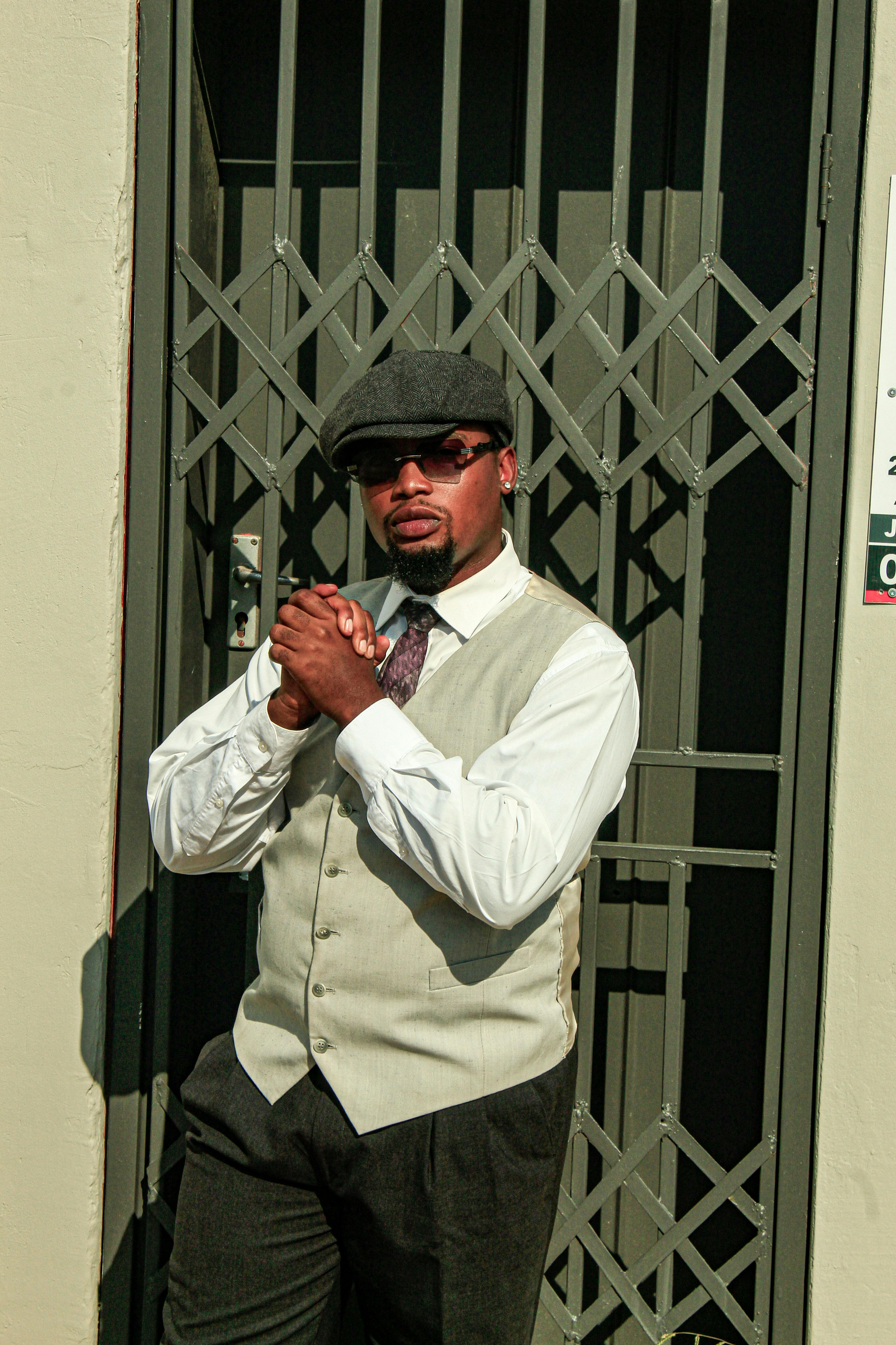 Man in vintage style outfit and cap posing outdoors against gate. Stylish fashion statement.