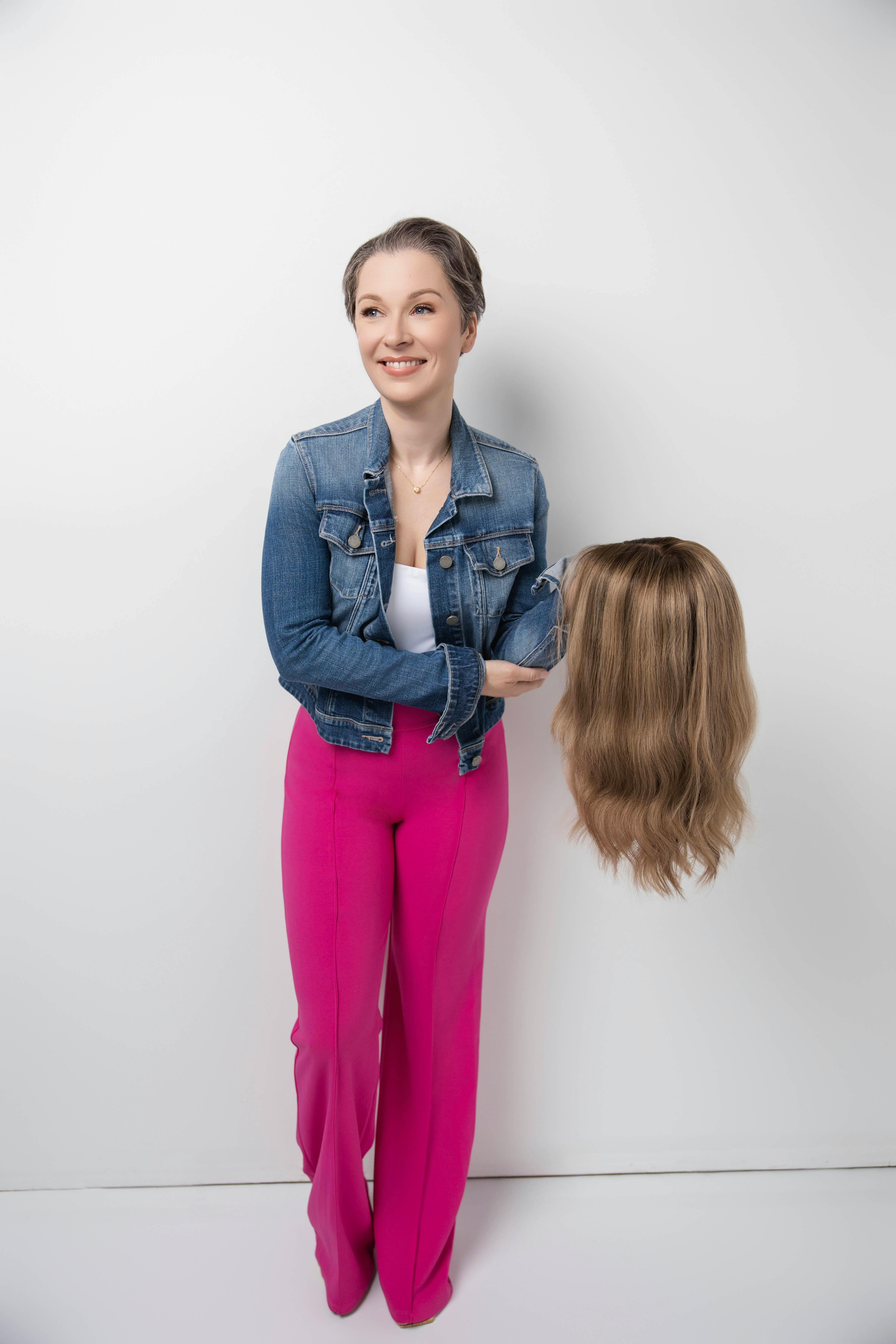 A smiling woman with alopecia holding a wig, showcasing confidence and style in a studio setting.