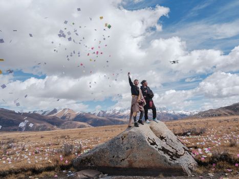 Two people on a rock, tossing confetti, with a drone hovering nearby in a mountainous landscape.
