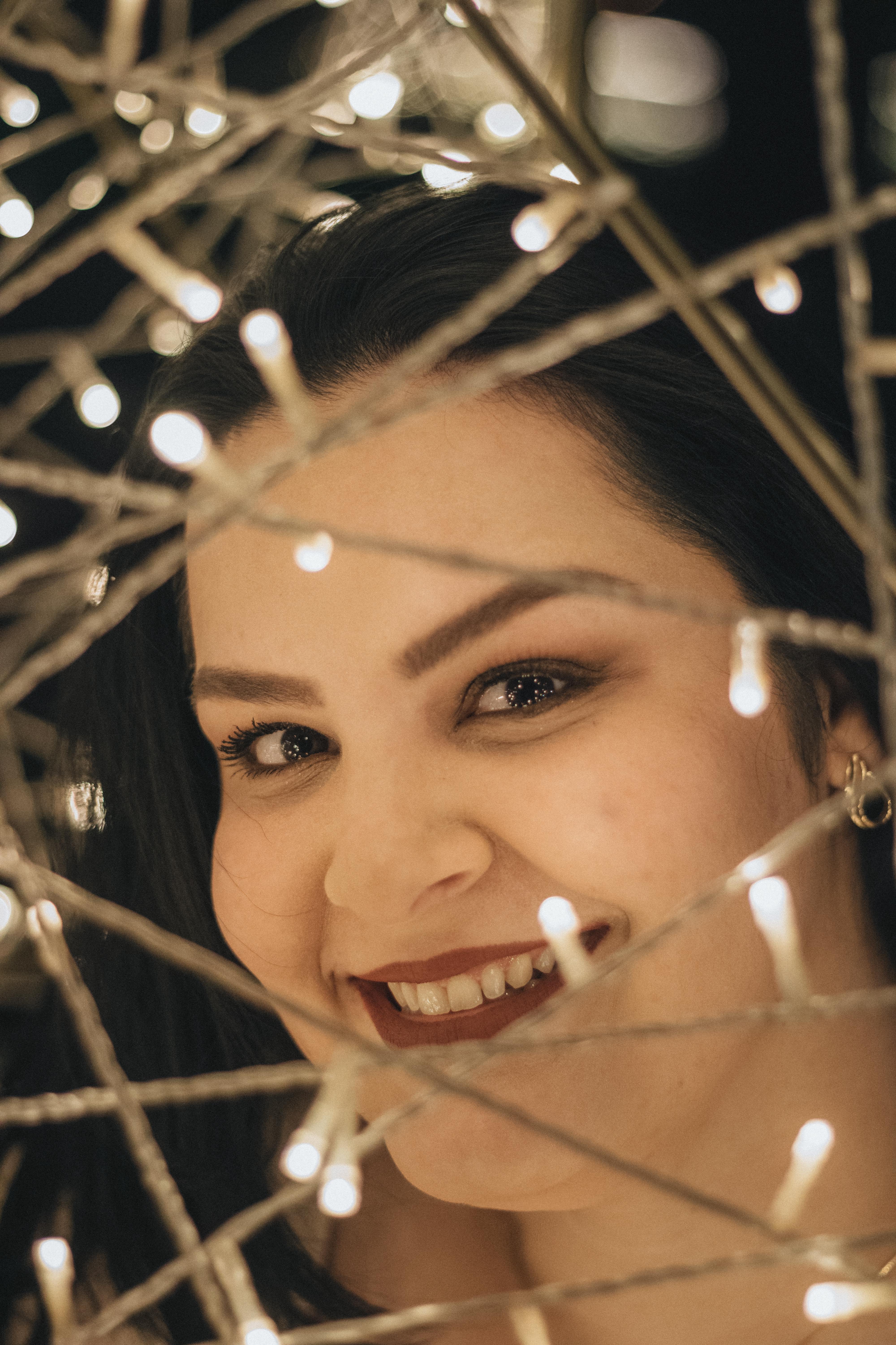 A joyful woman smiles through twinkling holiday lights, creating a warm festive ambiance.