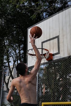 A male basketball player makes a jump shot on an outdoor court.