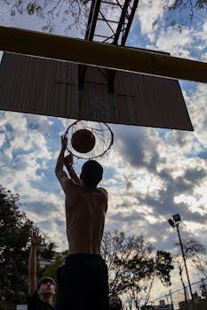 Silhouette of players in an outdoor basketball game with clouds in the background.