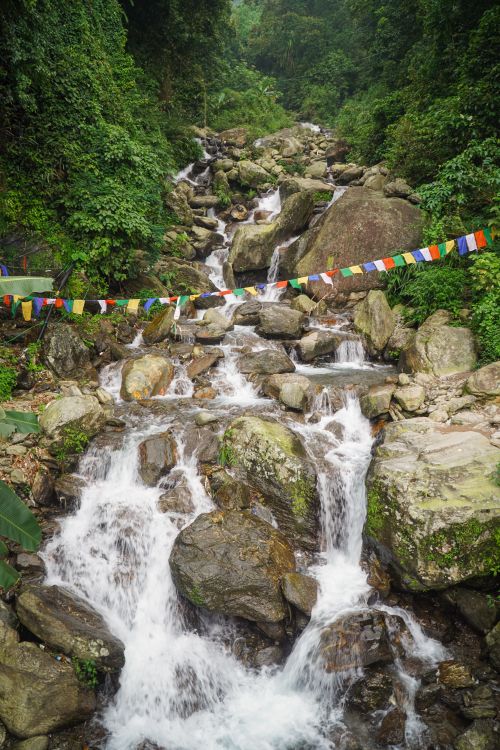 Free Scenic waterfall cascading through rocky terrain in Darjeeling, framed by vibrant prayer flags and green foliage. Stock Photo