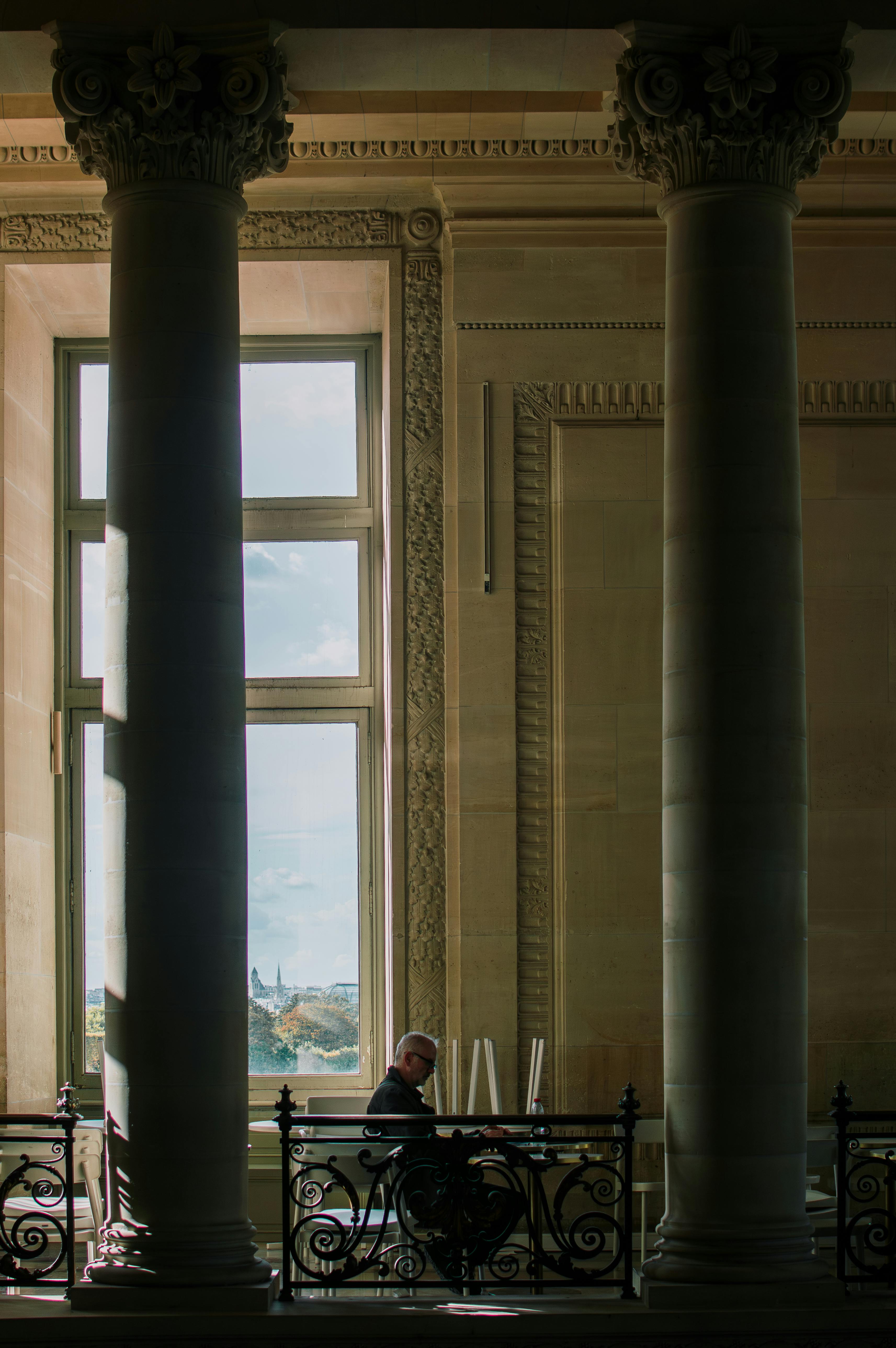 A man sits reading between grand columns in a historic Parisian building with a view of the city through large windows.