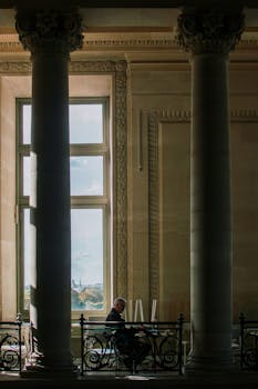 A man sits reading between grand columns in a historic Parisian building with a view of the city through large windows.