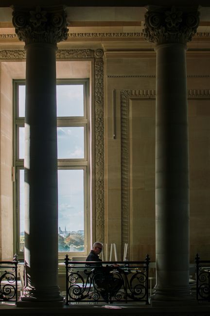 A man sits reading between grand columns in a historic Parisian building with a view of the city through large windows.