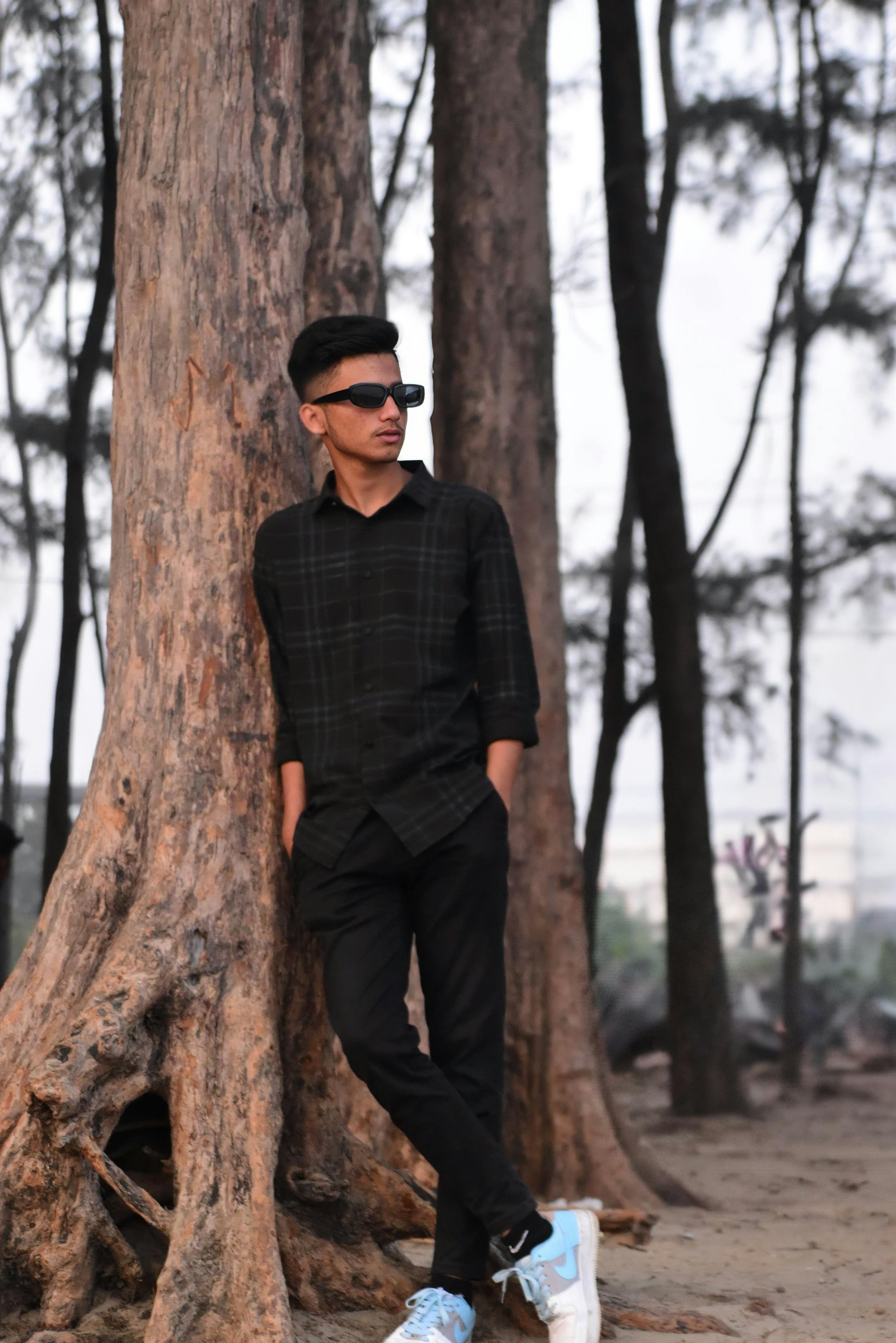Free Man in sunglasses and black attire leaned against a tree, creating a cool and stylish vibe. Stock Photo