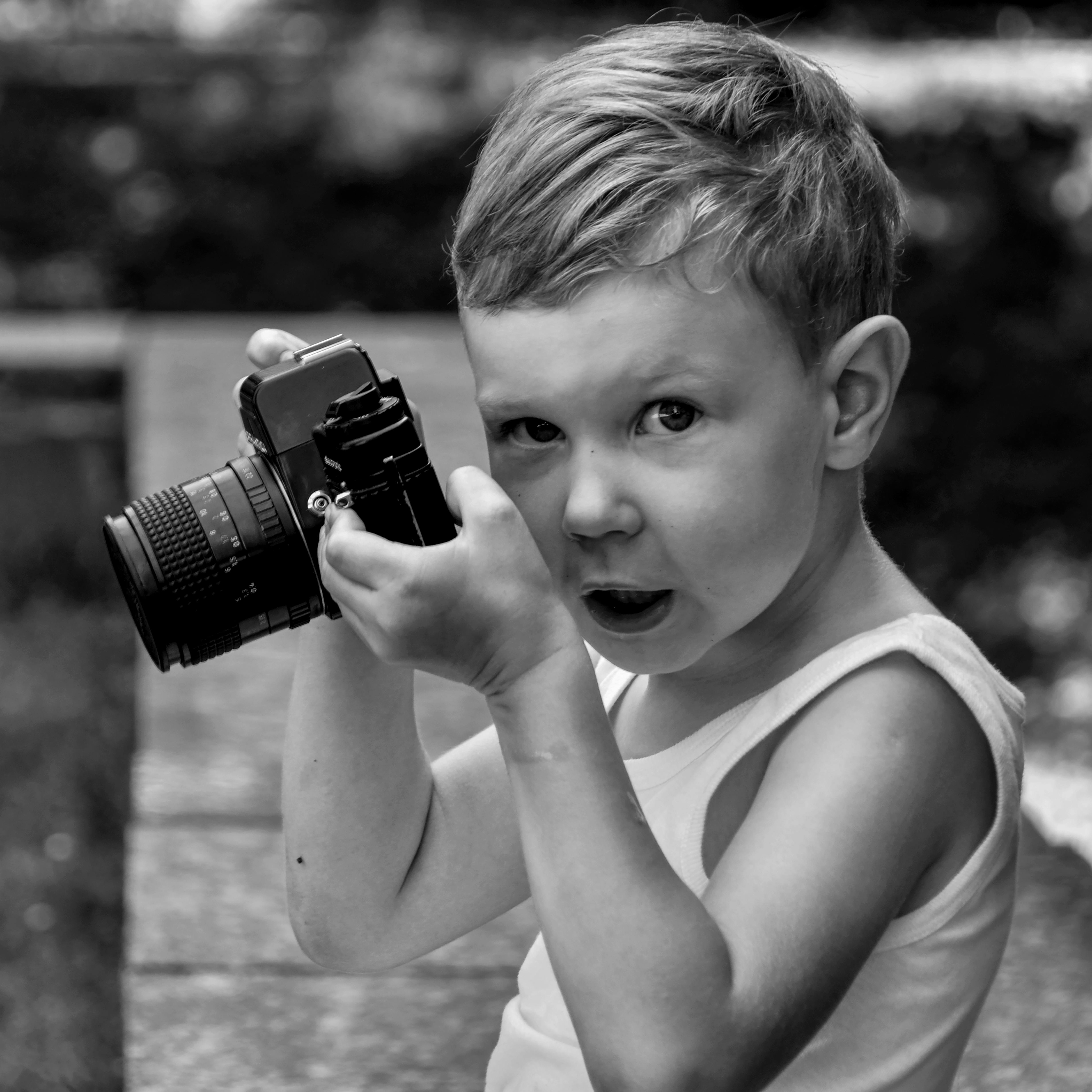 Black and white photo of a young boy holding a camera outdoors, capturing his curiosity.
