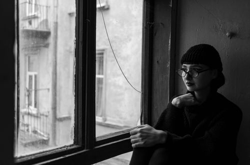 Free Black and white portrait of a woman in a beanie and glasses sitting by a window. Stock Photo
