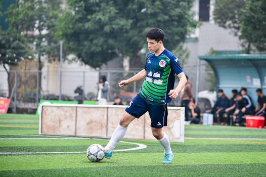 A soccer player in a green and blue uniform controls the ball on an outdoor field in Hà Nội, Việt Nam.