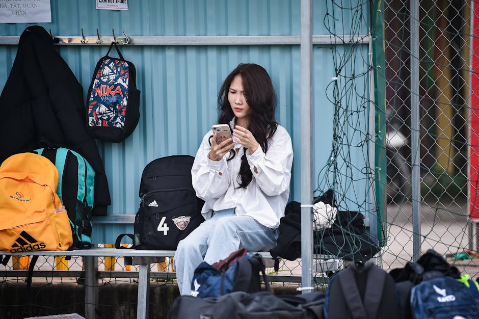 Young woman on a bench with backpacks, focused on her phone outdoors.