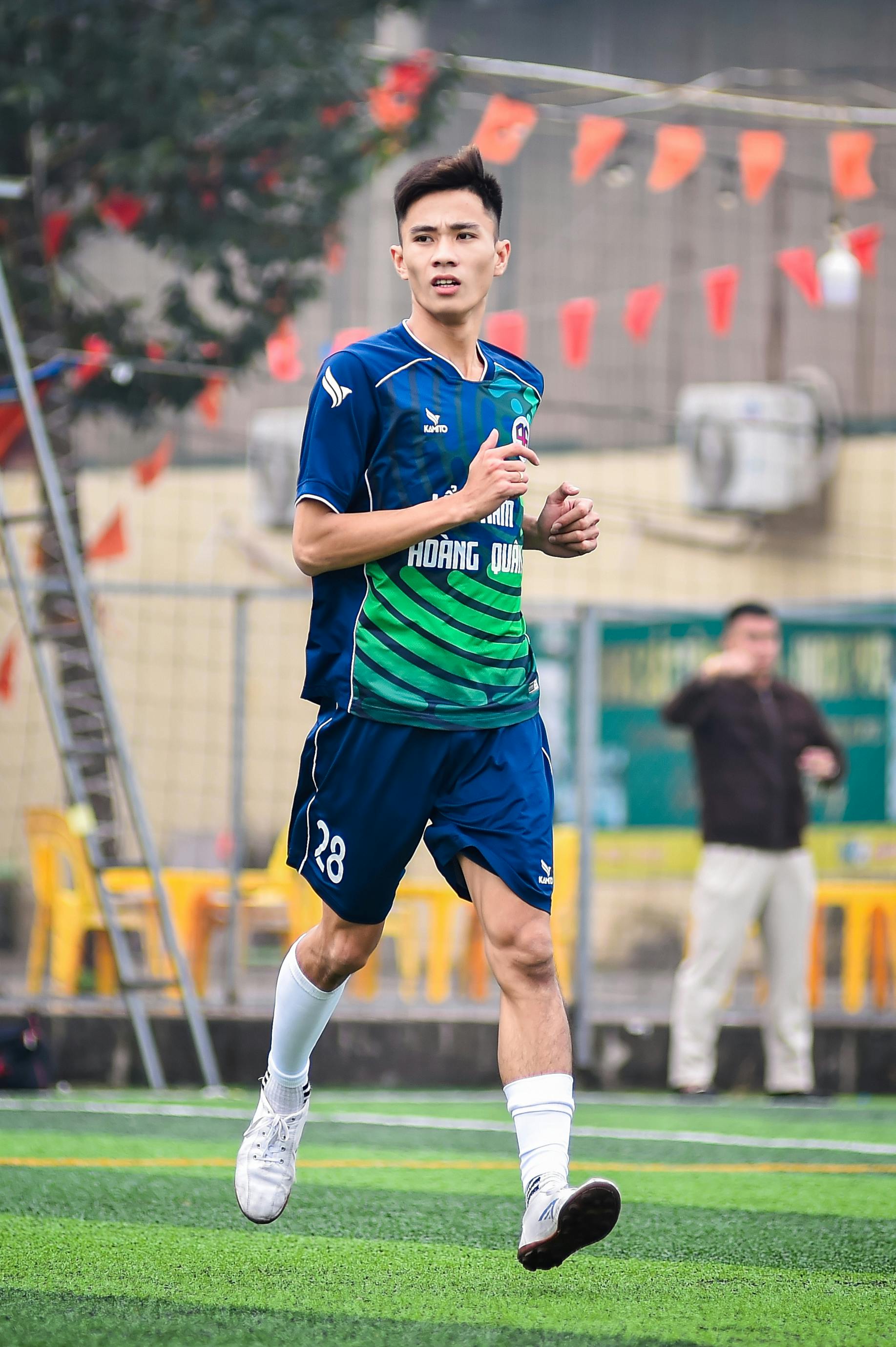 Asian footballer in vibrant kit jogging on a Hanoi soccer field during a daytime match.