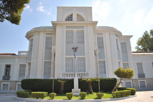 A grand historic building adorned with a Greek flag, surrounded by gardens.