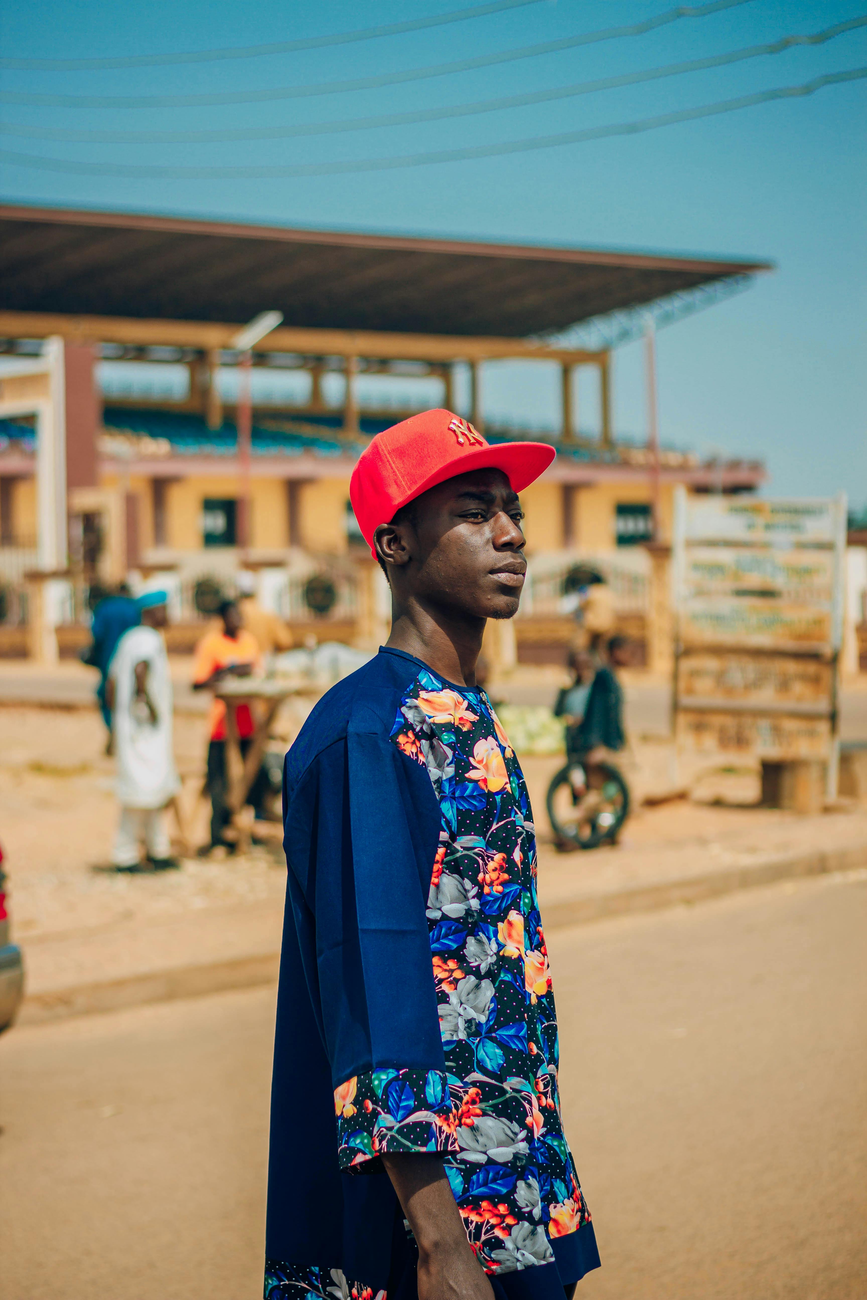 Portrait of a young man in striking attire with a red cap, standing on a lively street.