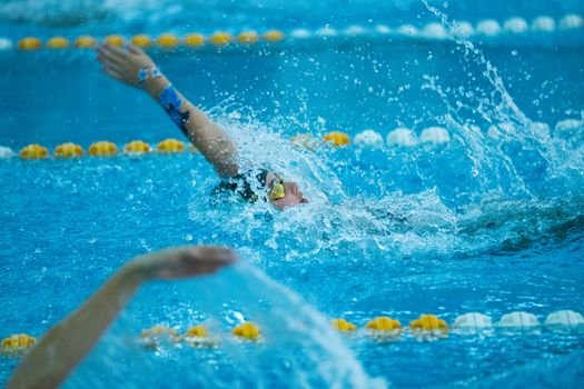 A competitive swimmer performs a backstroke in a pool, creating splashes.